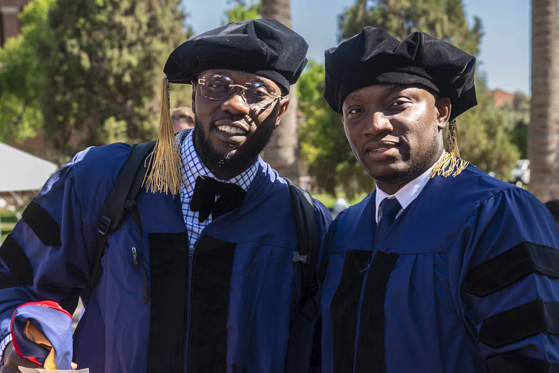 Two young men dressed in graduation regalia stand outside before convocation.