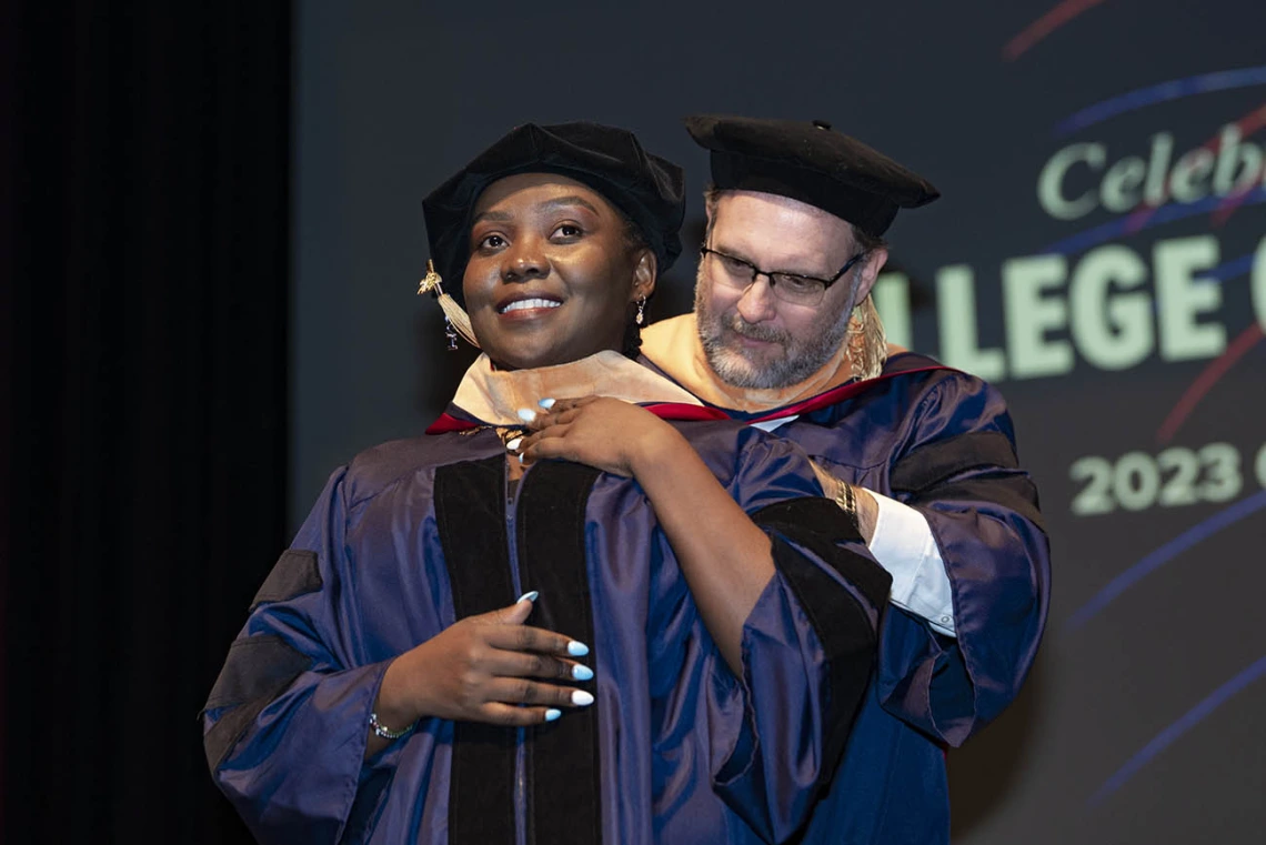 A young woman wearing graduation regalia smiles as she receives her hood by a male professor during convocation.