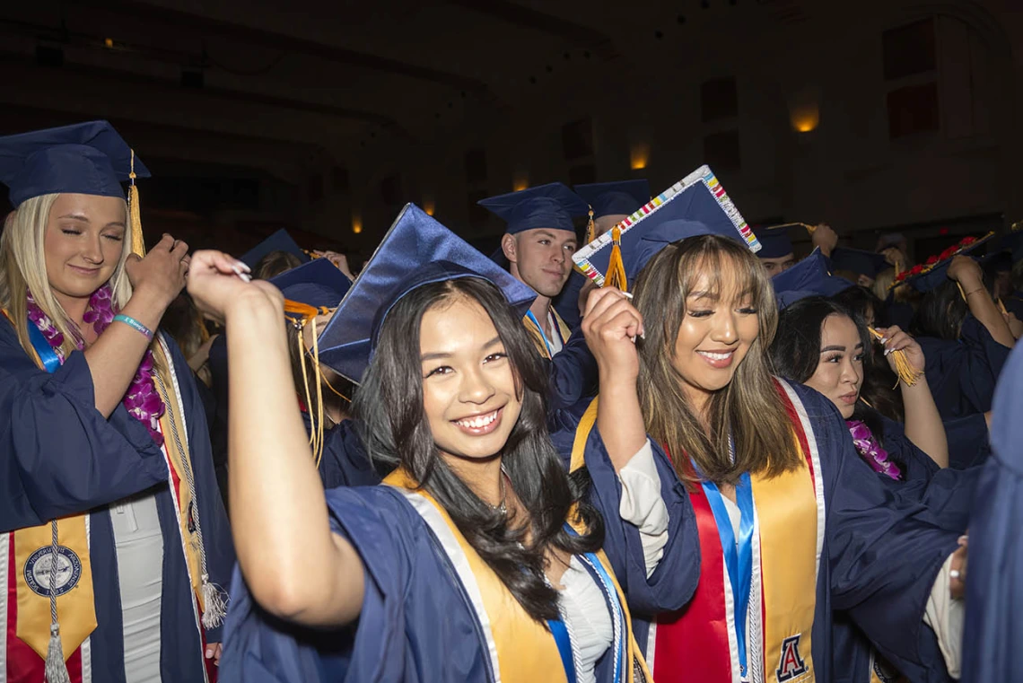 Two young women smile as they move their graduation cap tassle to the other side to signify their graduation.