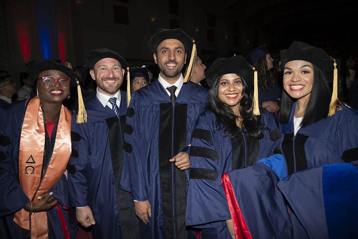 Five female and male PhD graduates in caps and gowns stand together smiling. 