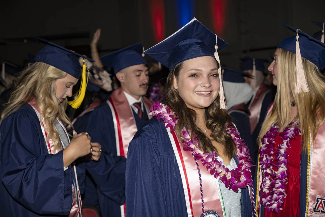 A smiling young woman in a graduation cap and gown stands in front of a larger group of graduates.