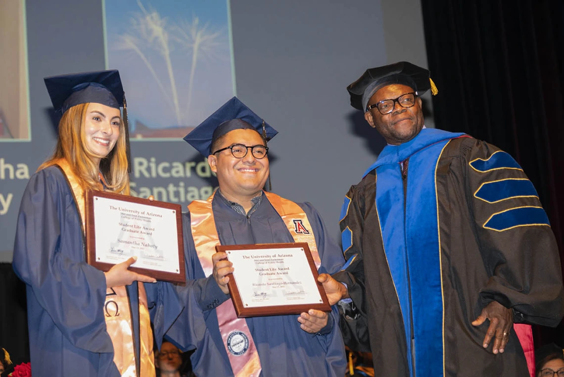 A female and male graduate in caps and gowns stand holding certificates next to a faculty member, also in graduation regalia. 