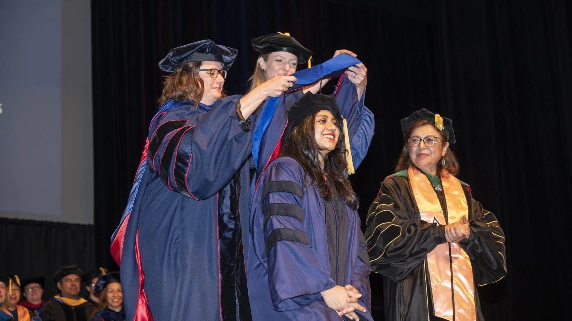 Two female faculty in graduation regalia place a sash over the shouldders of a smiling female graduate while another female faculty member looks on. 