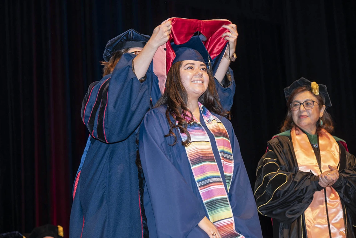 A smiling young woman in graduation cap and gown has a sash placed over her shoulders by a professor as the dean of the college looks on. 