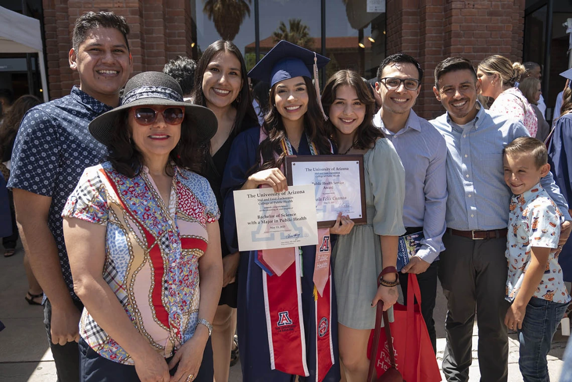 A family surrounds a young woman graduate in cap and gown holding two certificates. 