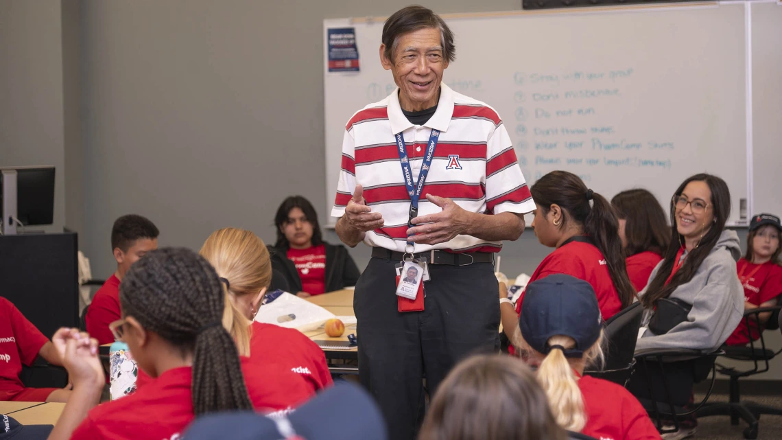 Older Asian man stands in a classroom setting surrounded by kids who are listening to him talk. 