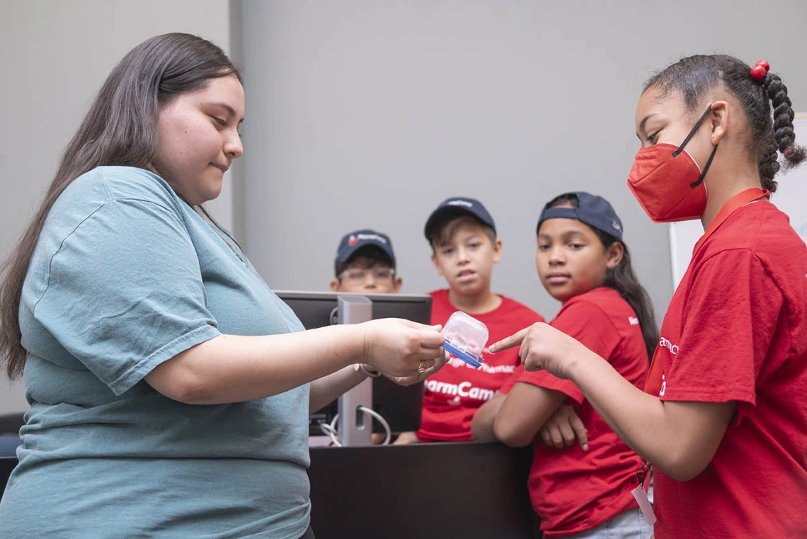 A young woman in blue shirt with long brown hair holds out a plastic container to a girl who is pointing at the container.