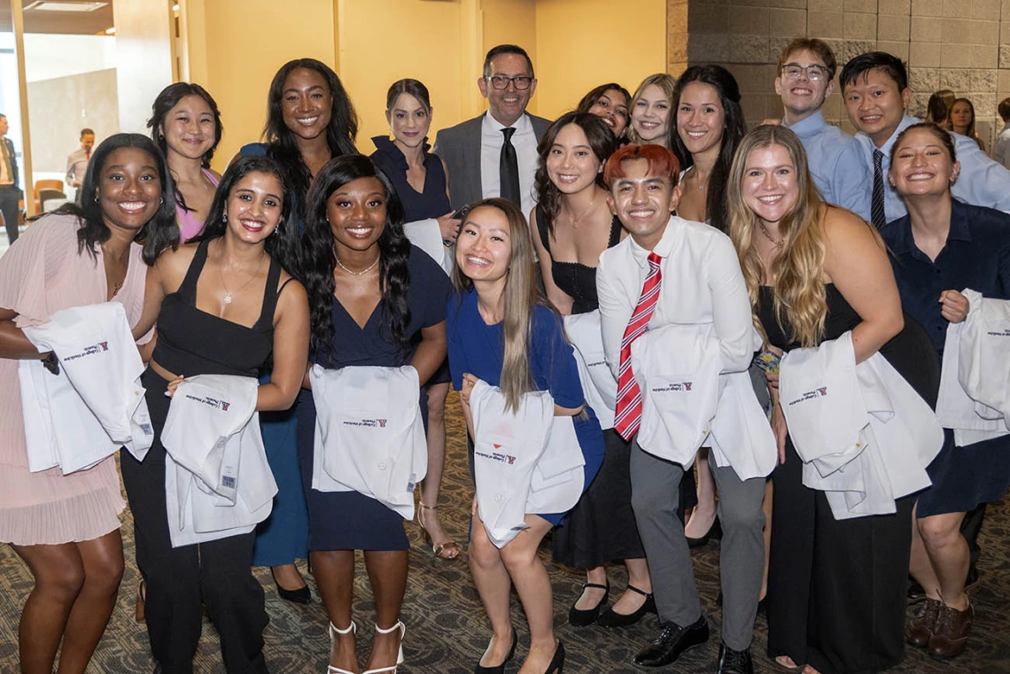 A group of about 16 medical students holding white coats over their arms pose for a photo with a male faculty member. All are smiling. 