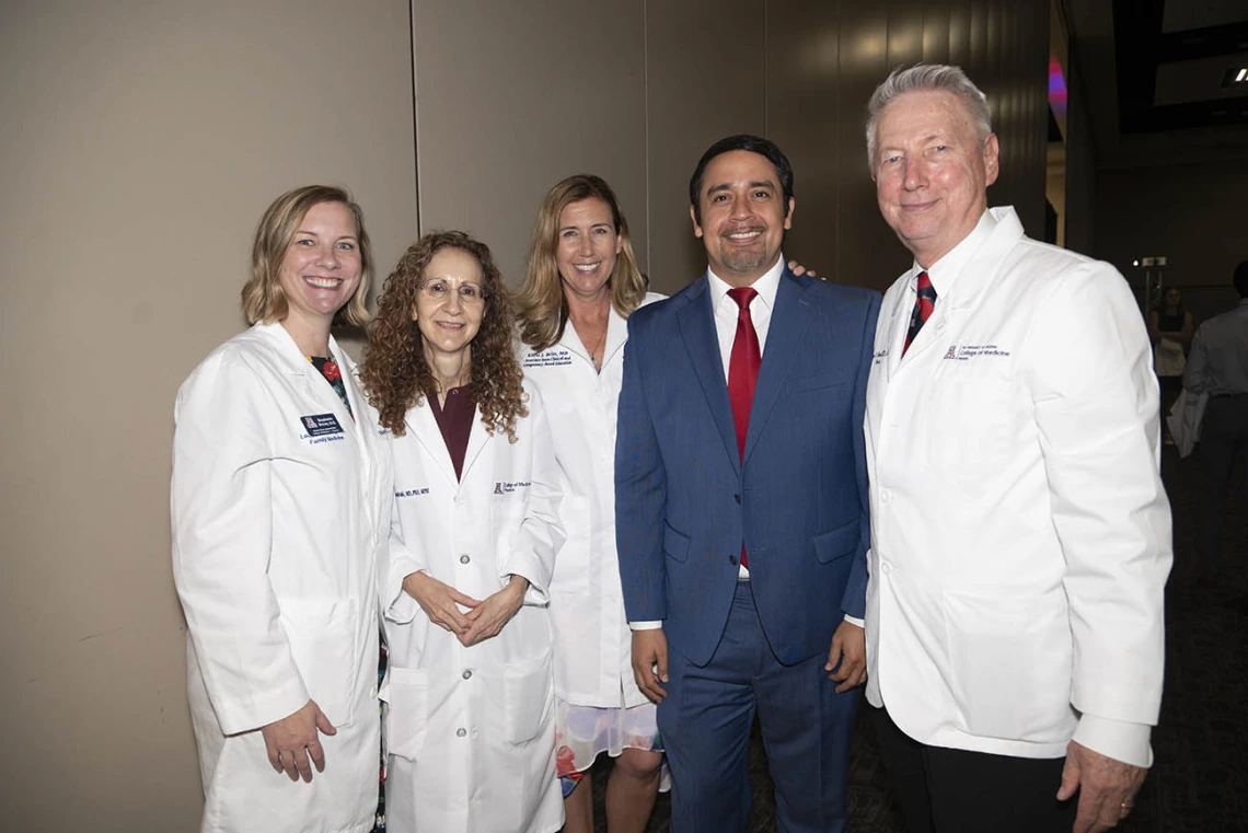 Five medical school faculty members, women and men, mostly wearing white coats stand together smiling. 