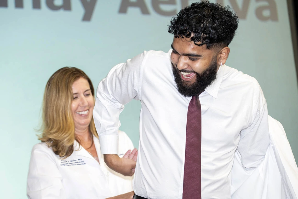 A white female doctor helps a young male medical student with brown skin and a beard put on his white coat. 