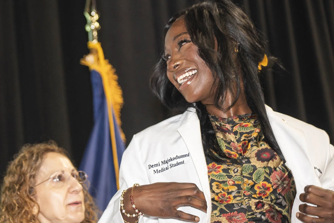 A white female doctor helps a young female medical student with dark skin and a big smile put on her white coat.