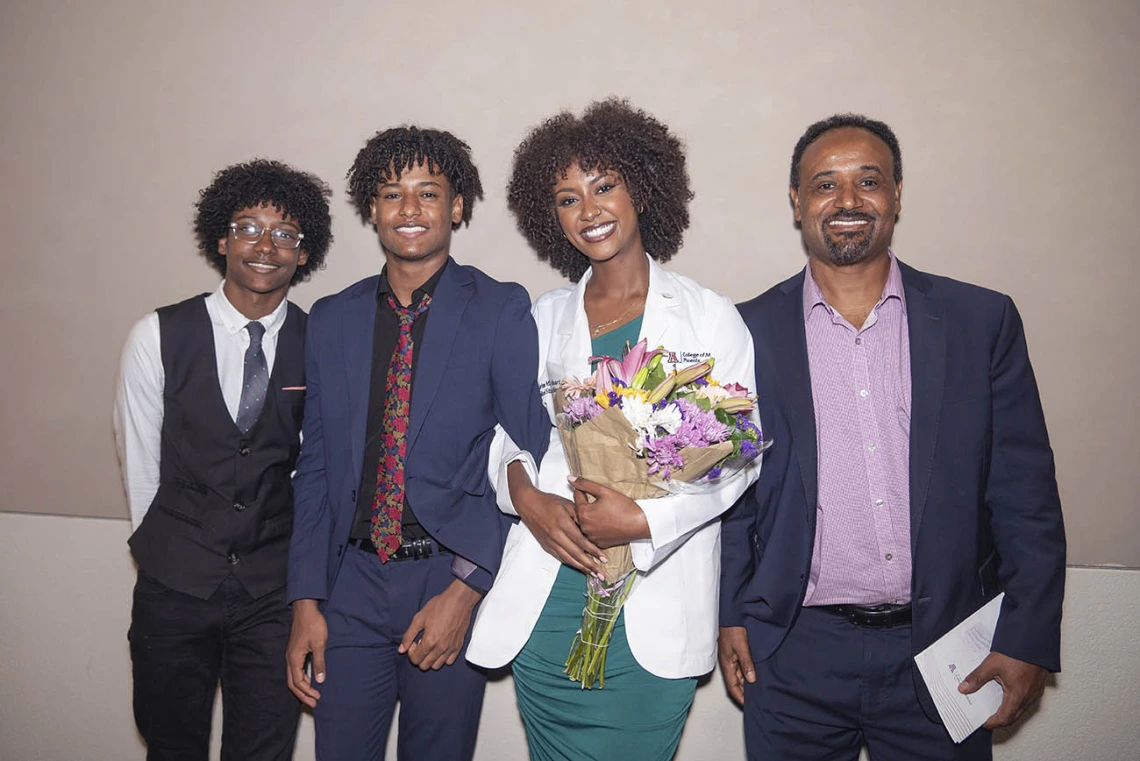 A young female medical student with dark skin smiles holding flowers as she stands withher family.