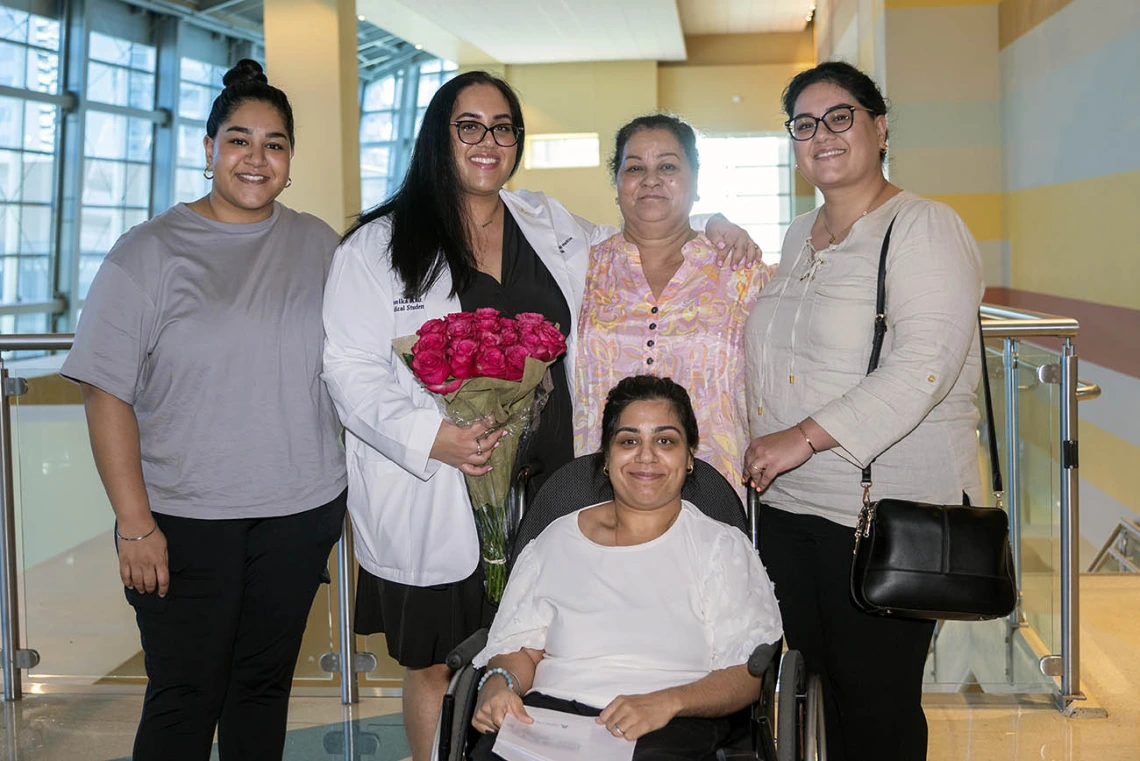 A young woman in a white coat holding flowers and smiling stands with four female members of her family.