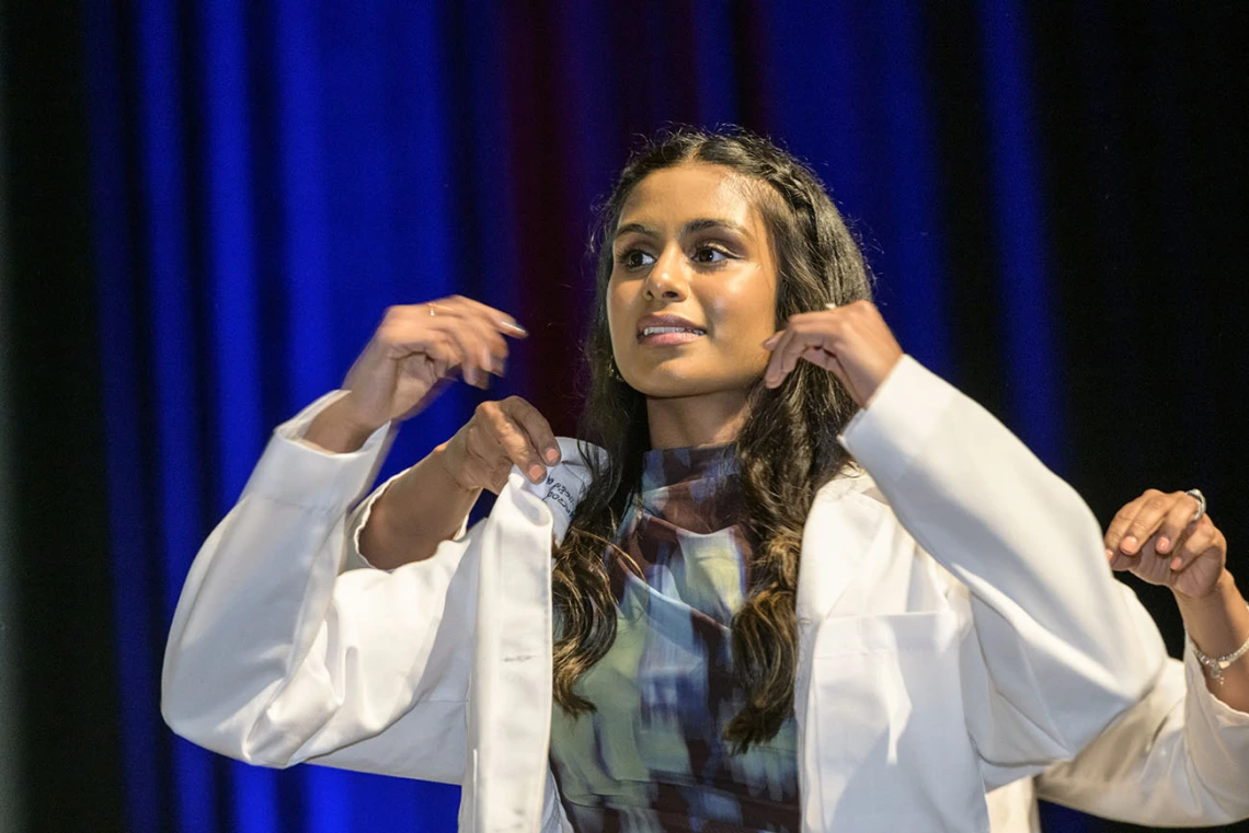 A young, brown-skinned femal medical student with long dark hair looks puts on her white medical coat.