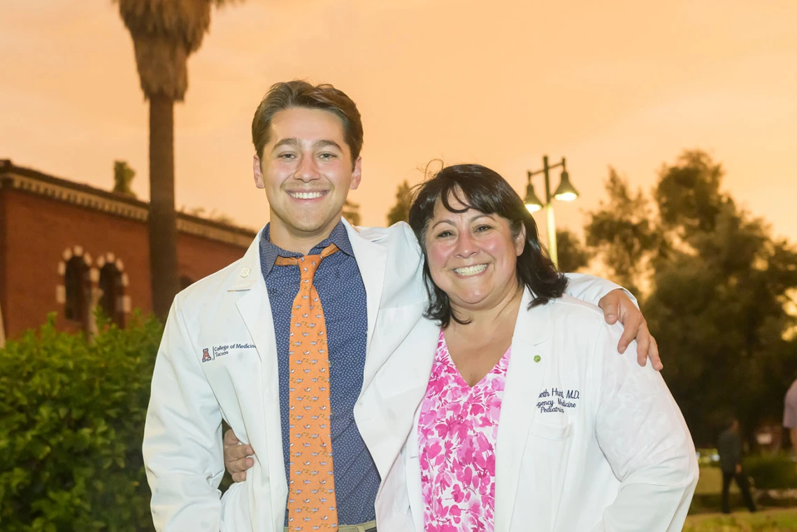 A young white male medical student stands with his arm around his mom, both are wearing white medical coats and smiling. 