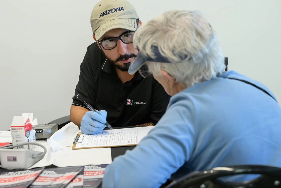 A man with a dark goatee, glasses and a baseball cap wearing medical gloves fills out paperwork for a woman with short gray hair. 