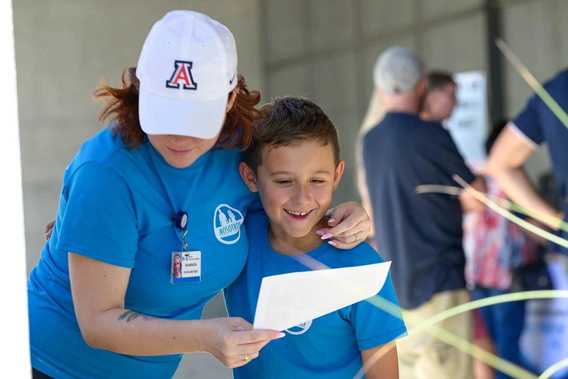 A woman with a white Arizona baseball hat and bright blue shirt has her arm around her son while showing him a piece of paper. 