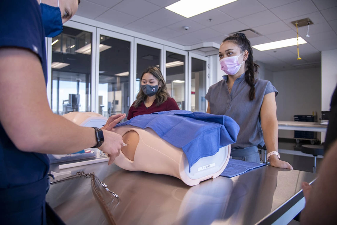 AGACNP student Katie Pon Rambo, in gray scrubs, watches as another student practices inserting a chest tube. This training is required for graduates to apply for credentials post-graduation.  