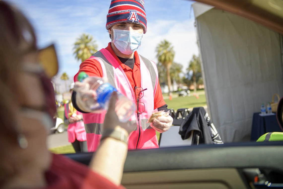 UArizona employee Brandon Goldstein hands out bottles of water and crackers for those waiting in an observation lot after being vaccinated.