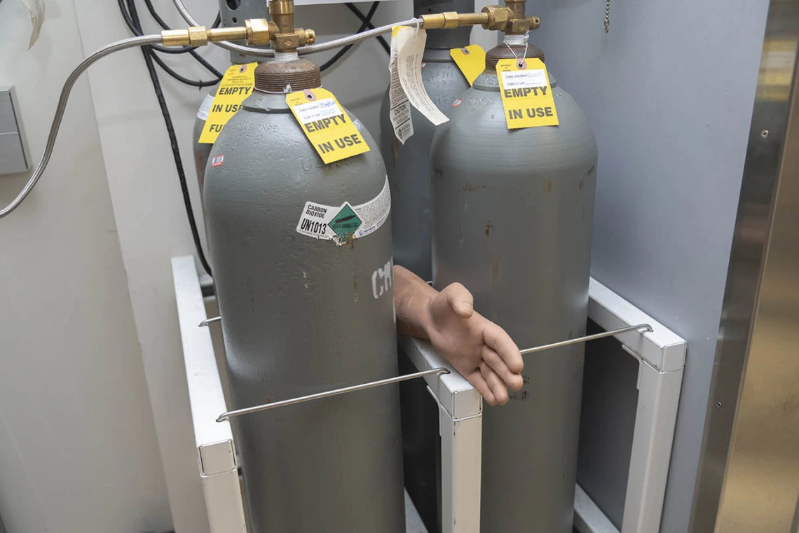 A hand sticks out from between two larg metal tanks with warning labels on them in a lab setting. 