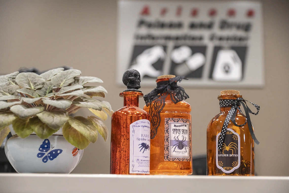 Three orange jars sit on a desk with labels that represent types of poisons. 