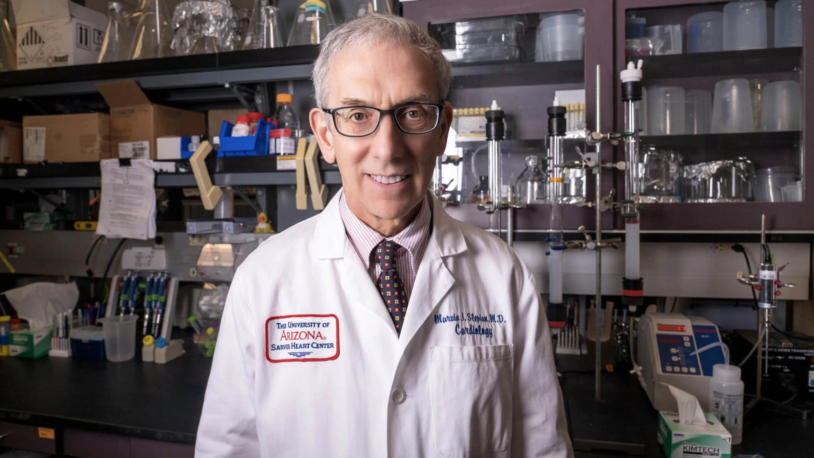 Older man with glasses and white hair stands smiling in a lab wearing a white coat.