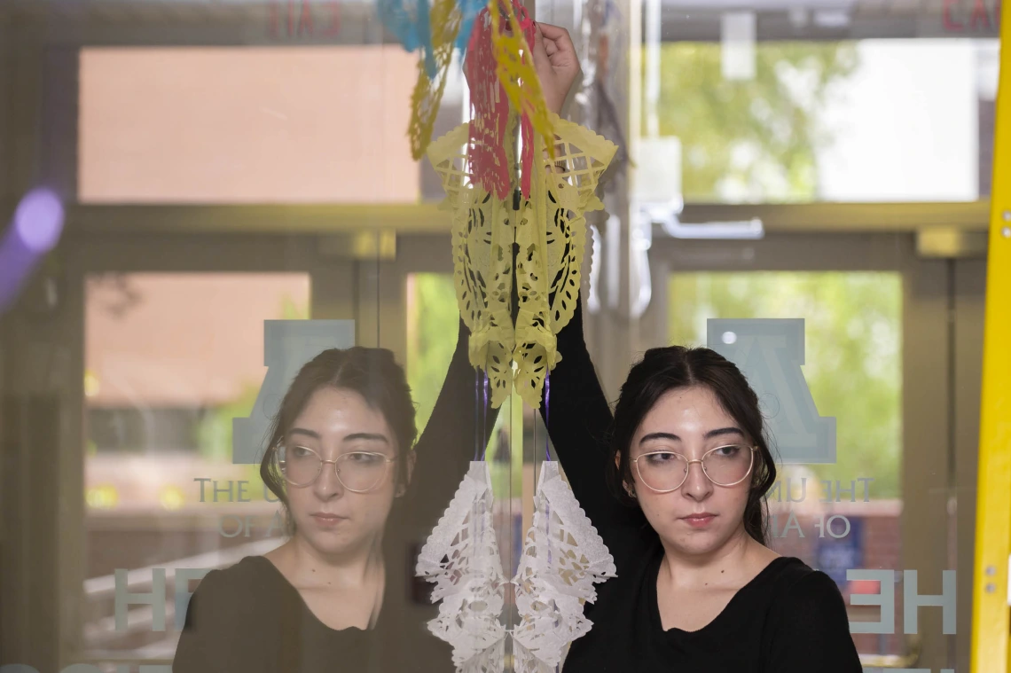 A young woman with long black hair holds up a tissue paper decoration against a window with her mirrored reflection. 