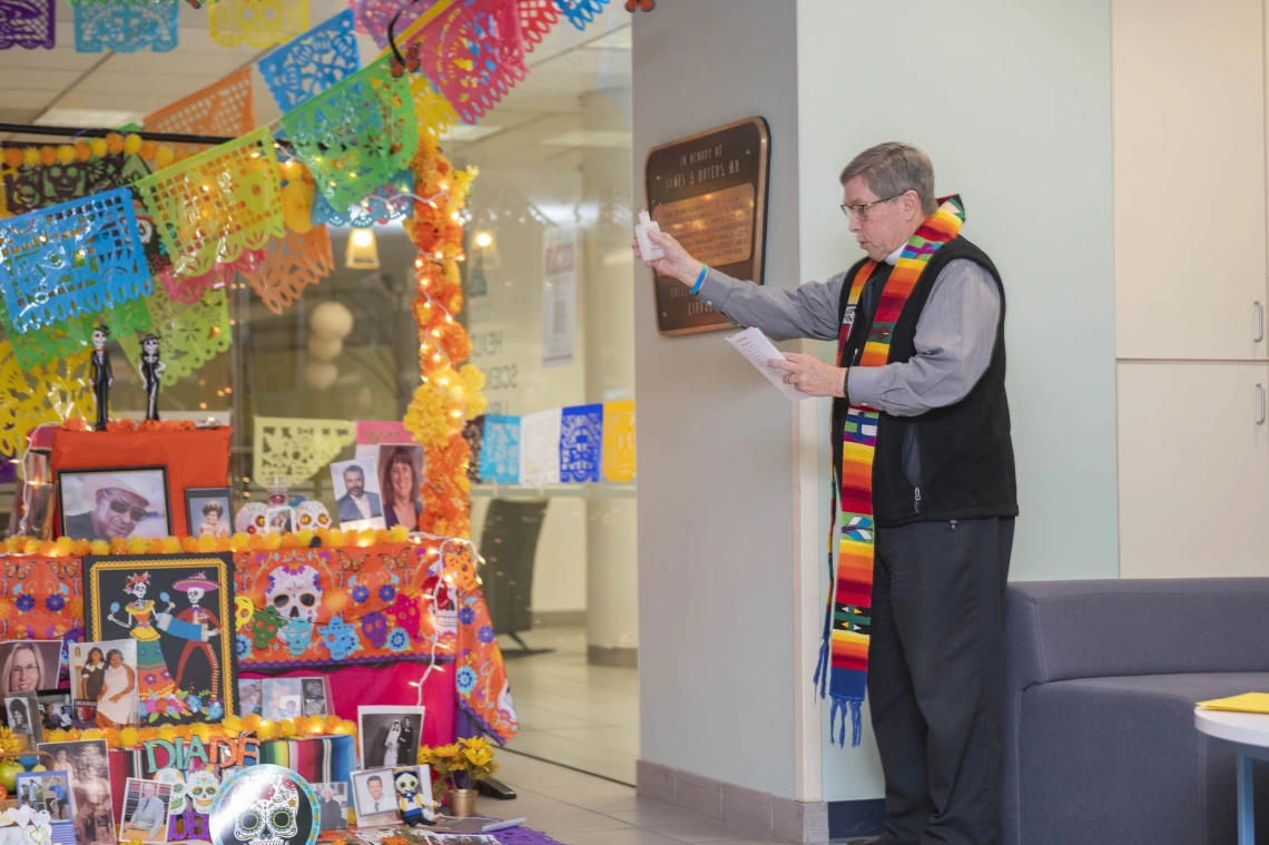 Chaplain Joe Fitzgerald stands beside the colorful Dia de los Muertos altar with his hand raised as he reads a blessing. 