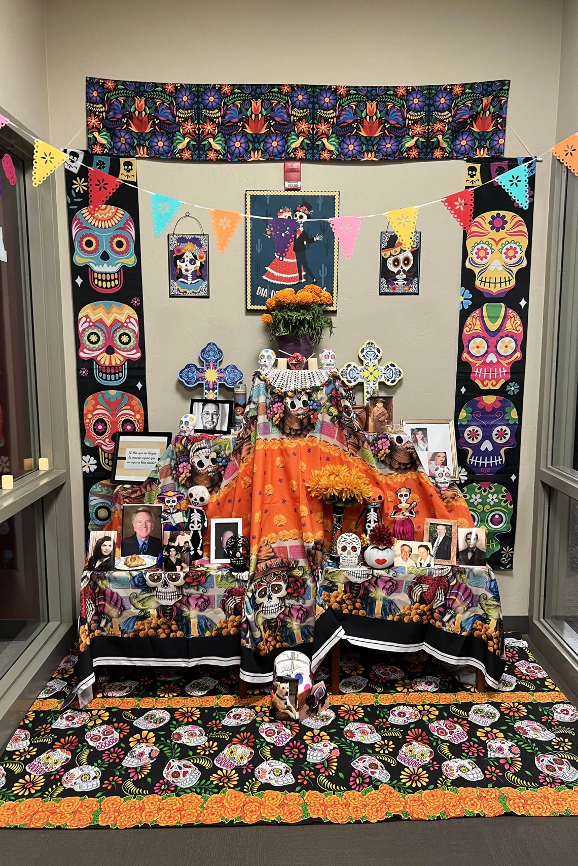 Brightly colored Dia de los Muertos altar with colorful skull design and many photographs on the table. 