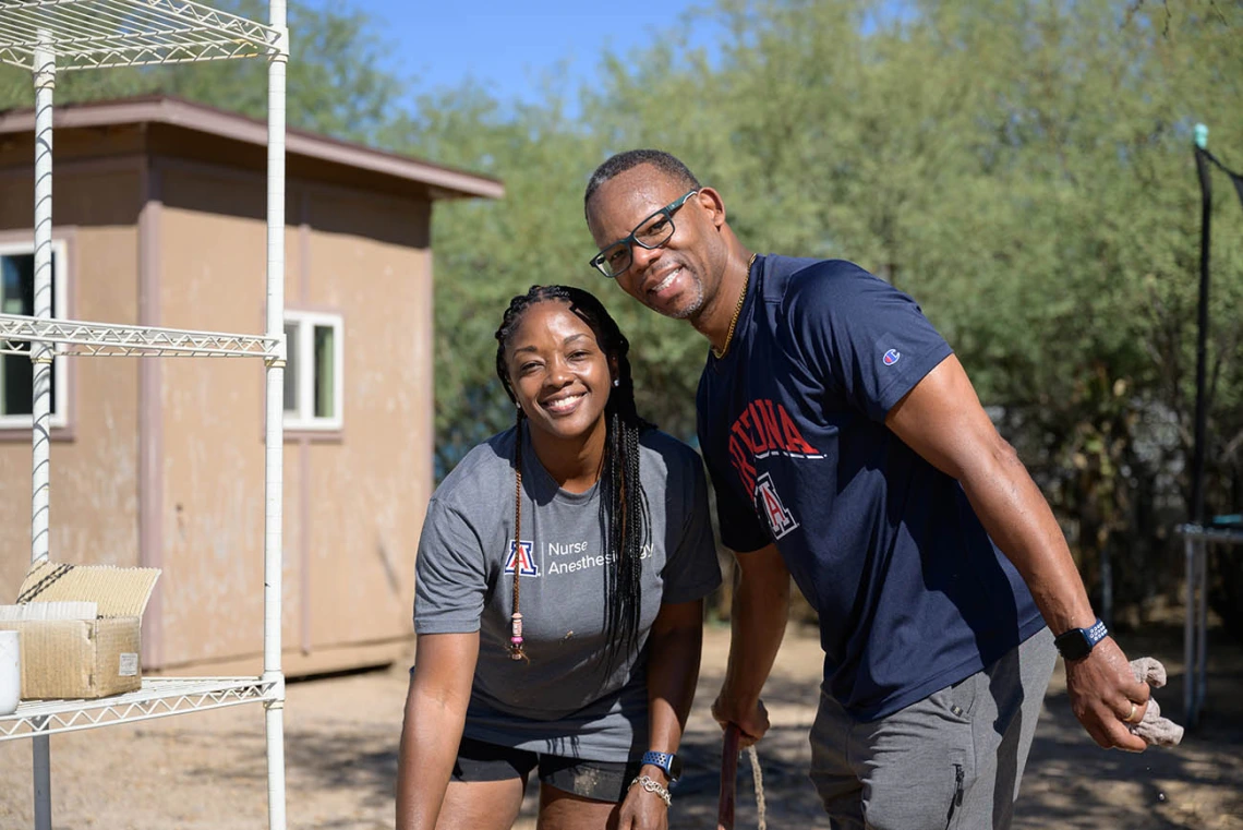 A young black woman and man stand next to each other smiling as they pause from volunteer work outside a small building. 