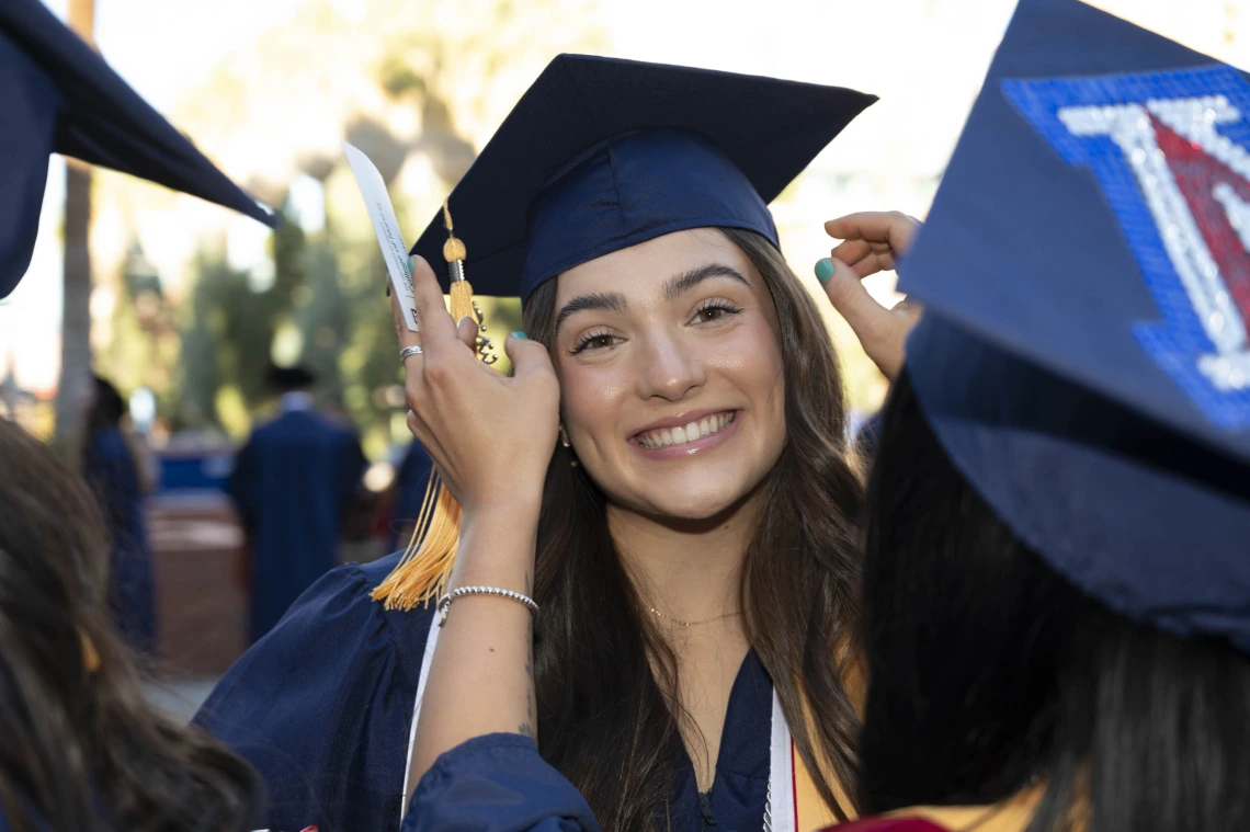 A smiling young woman wearing a graduation cap and gown has her hair adjusted by a friend. 