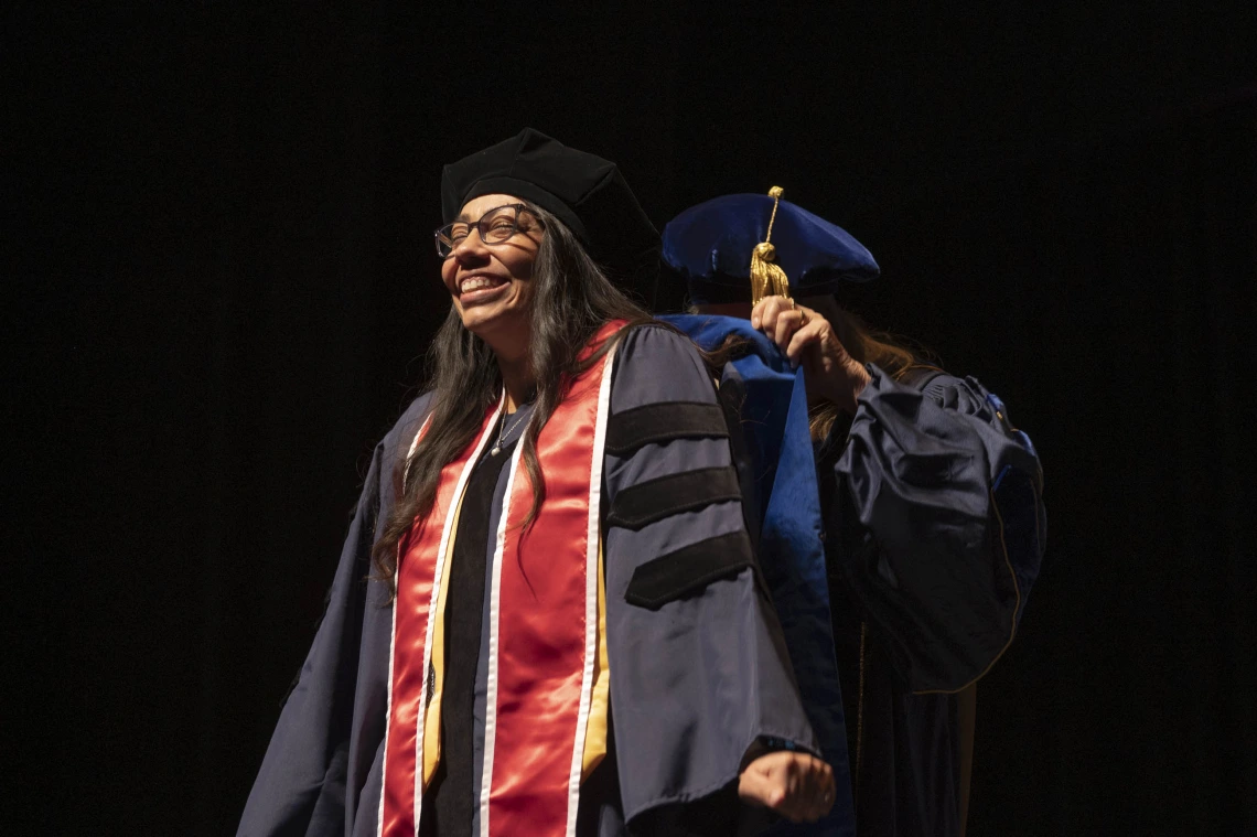 A nursing PhD candidate stands with a big smile on her face as a professor puts her graduation hood on her. Both are in caps and gowns. 