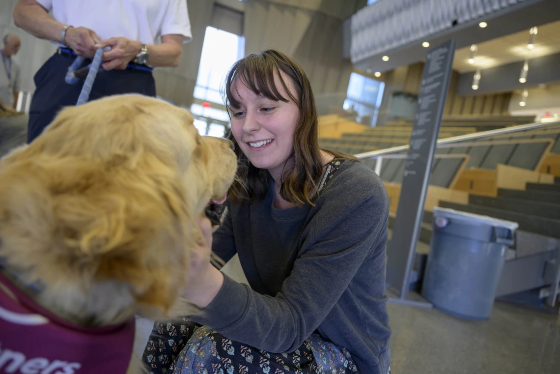 A woman kneels down looking into the face of a golden retriever while petting it. 