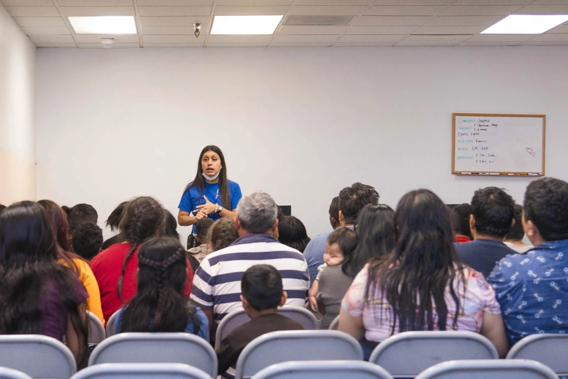 A woman in a blue shirt speaks to a crowd of migrants of all ages seated in metal folding chairs in a plain room.