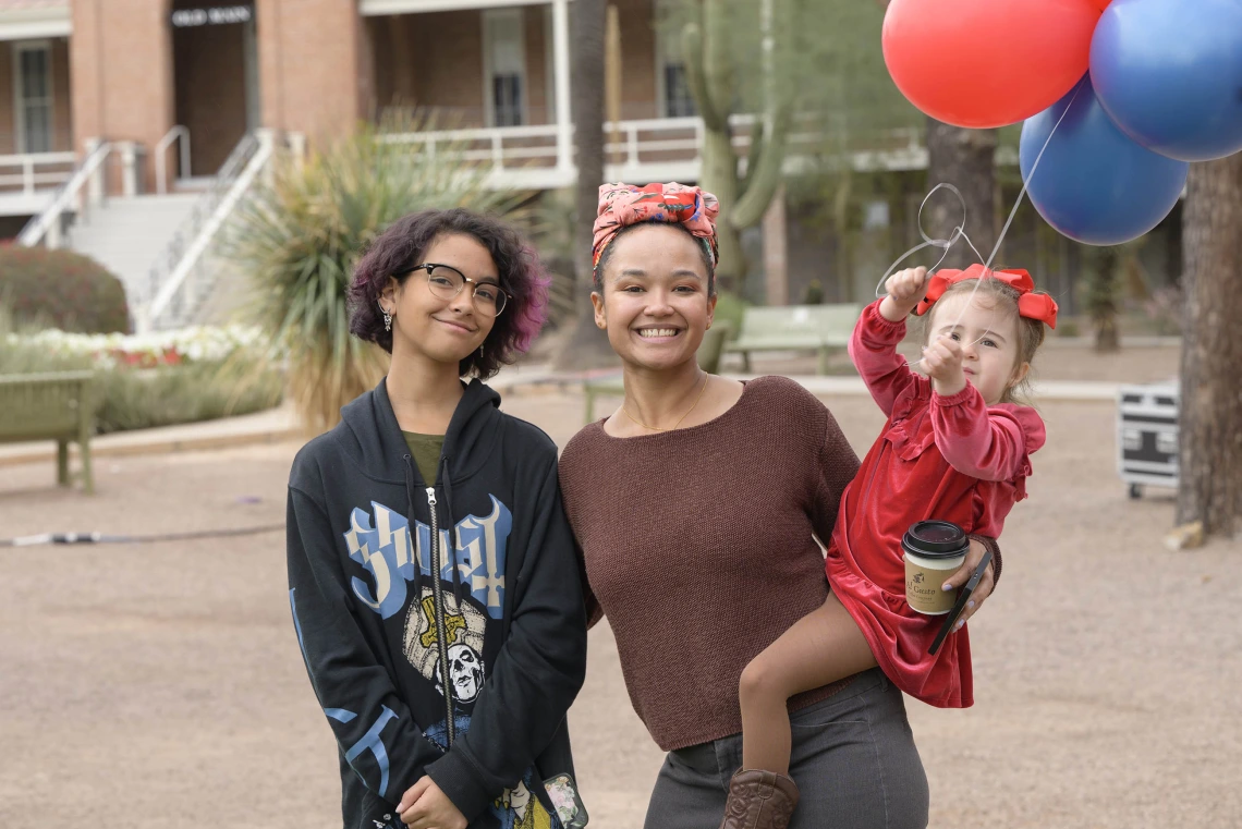 Two women stand next to each other, smiling. One is holding a small child who is holding balloons.