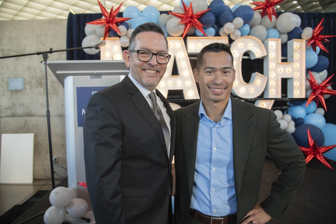 Two men in suitcoats smile as they stand in front of a stage with balloons at the University of Arizona College of Medicine – Phoenix Match Day event.