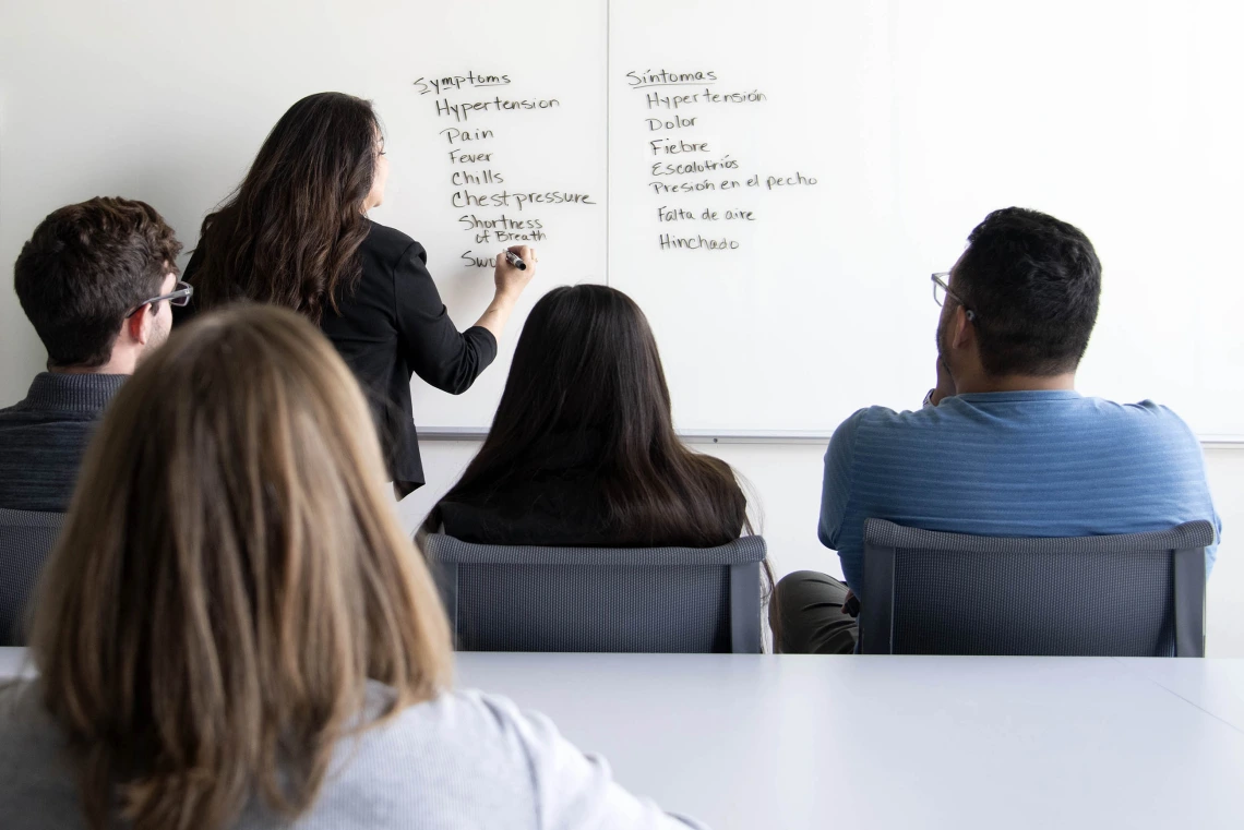 Students observe an instructor writing a list of medical terms in English and Spanish on a whiteboard