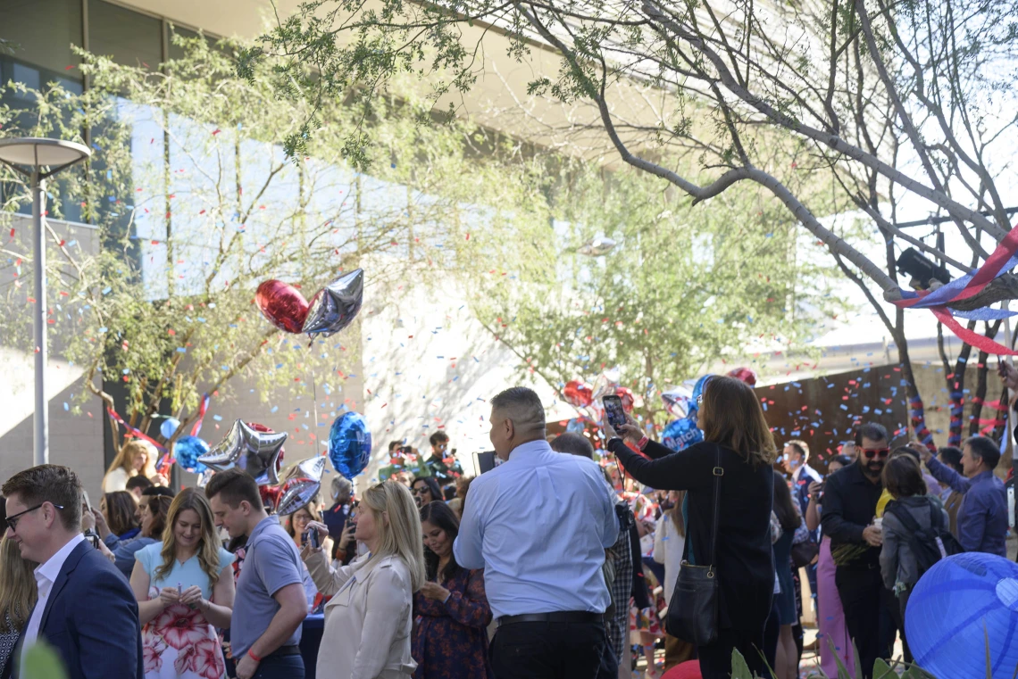 A crowd of people stands outside with confetti flying in the air. Lots of balloons decorate the space. 