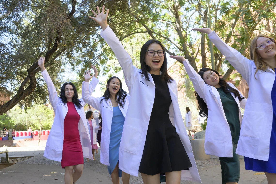 Several University of Arizona R. Ken Coit College of Pharmacy students wearing white coats raise their hands and cheer as they walk outside. 