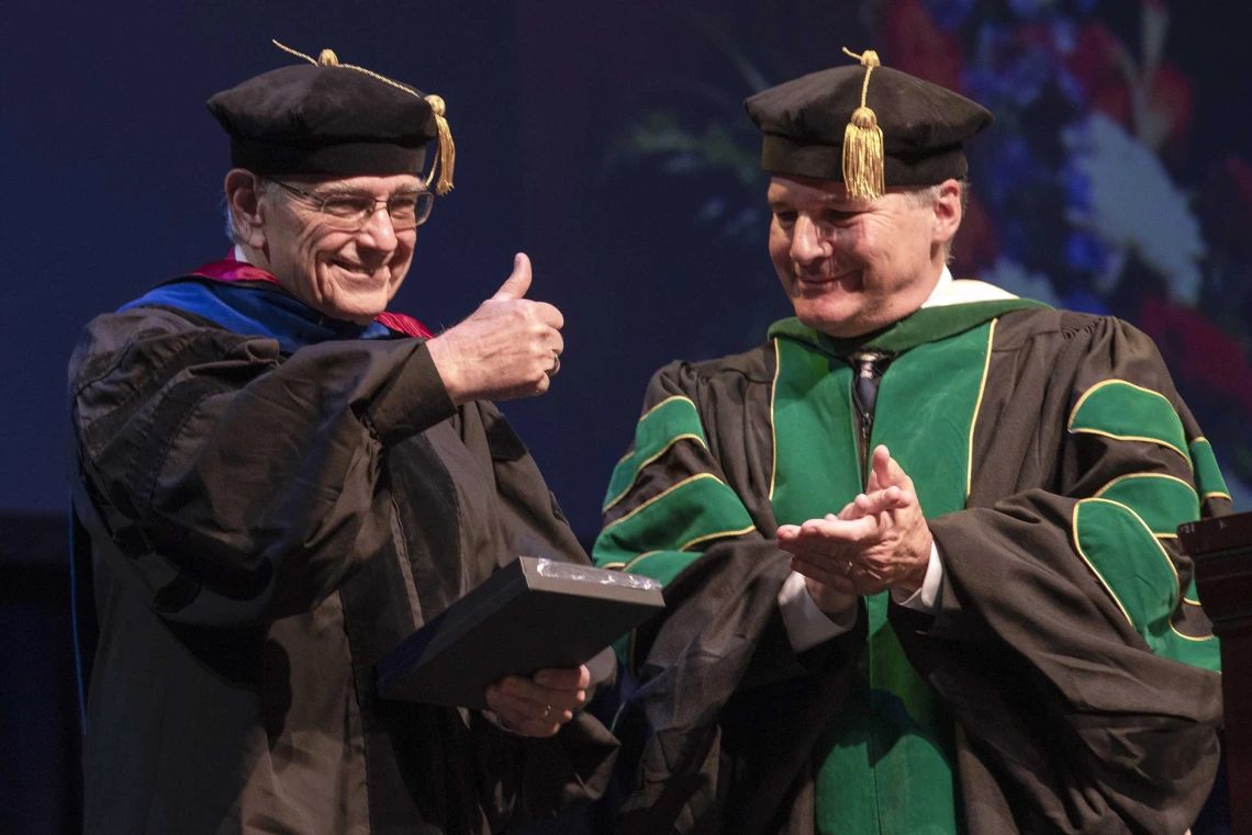 Two professors dressed in graduation regalia stand next to each other smiling as one hods a plaque and gives a thumbs-up. 