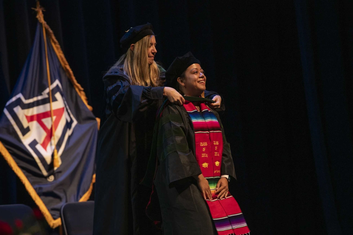 A University of Arizona College of Medicine – Phoenix student in graduation regalia smiles as a professor places a graduation hood on her.