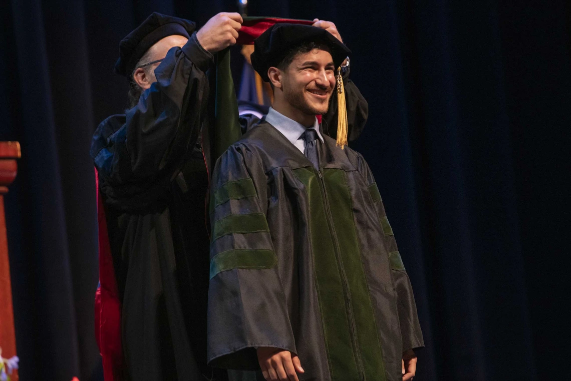 A University of Arizona College of Medicine – Phoenix student in graduation regalia smiles as a professor places a graduation hood on him. 