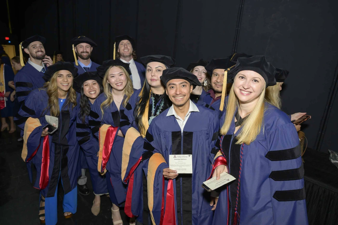 About a dozen smiling University of Arizona College of Nursing students wearing graduation caps and gowns stand backstage waiting for their convocation ceremony to begin. 