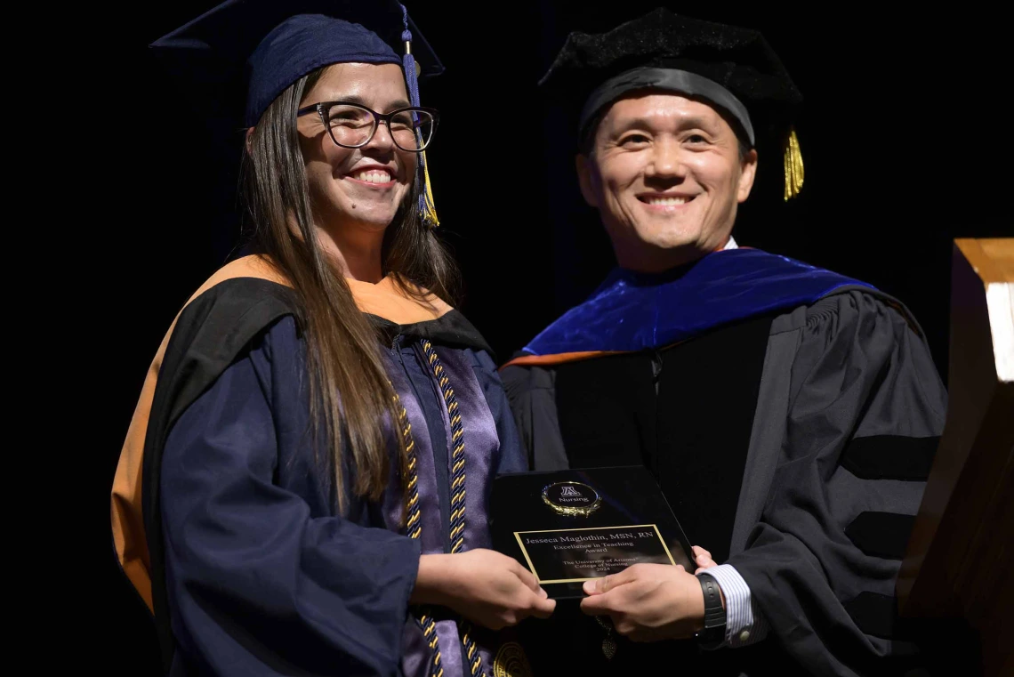 Two University of Arizona College of Nursing faculty members in graduation caps and gowns stand together smiling as they hold an award plaque in their hands. 