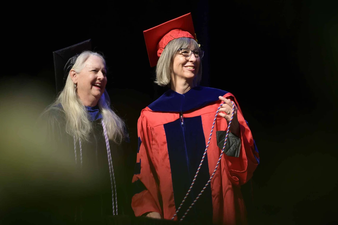 Two University of Arizona College of Nursing faculty dressed in graduation caps and gowns stand on a stage smiling as they look off to the side. 