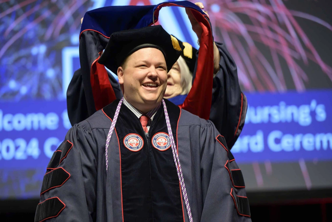 A University of Arizona College of Nursing professor places a hood over the shoulders of a nursing student. Both are dressed in graduation caps and gowns.