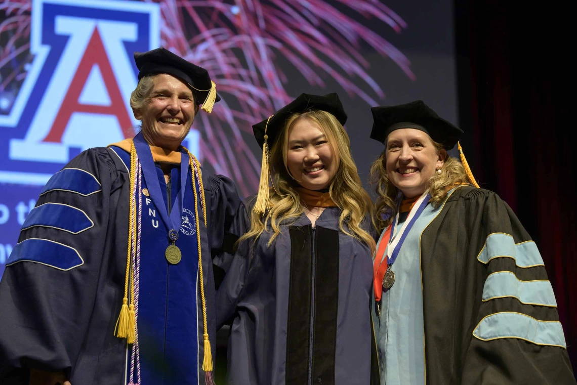 Two University of Arizona College of Nursing professor flank a student on stage. All are wearing graduation caps and gowns. 