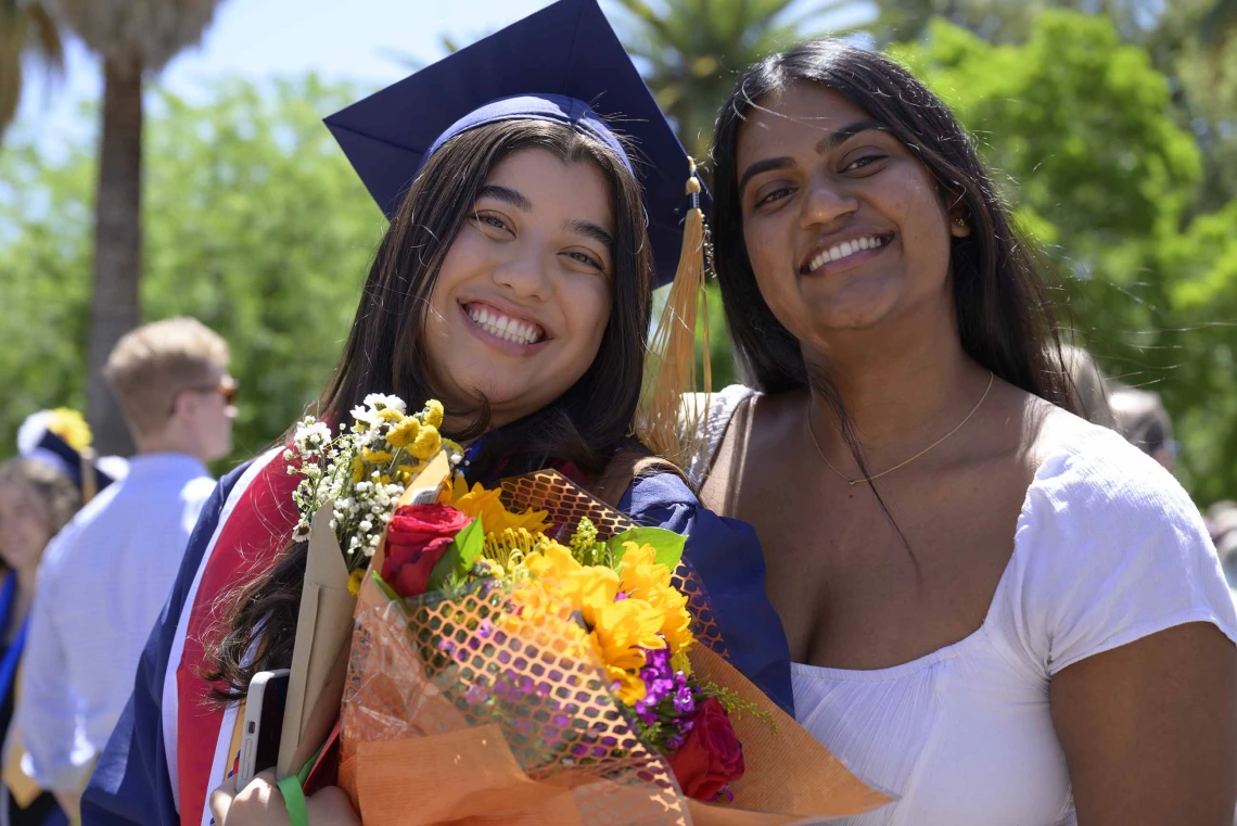 A University of Arizona College of Nursing student in a cap and gown holding flowers stands next to her friend outside. Both are smiling. 