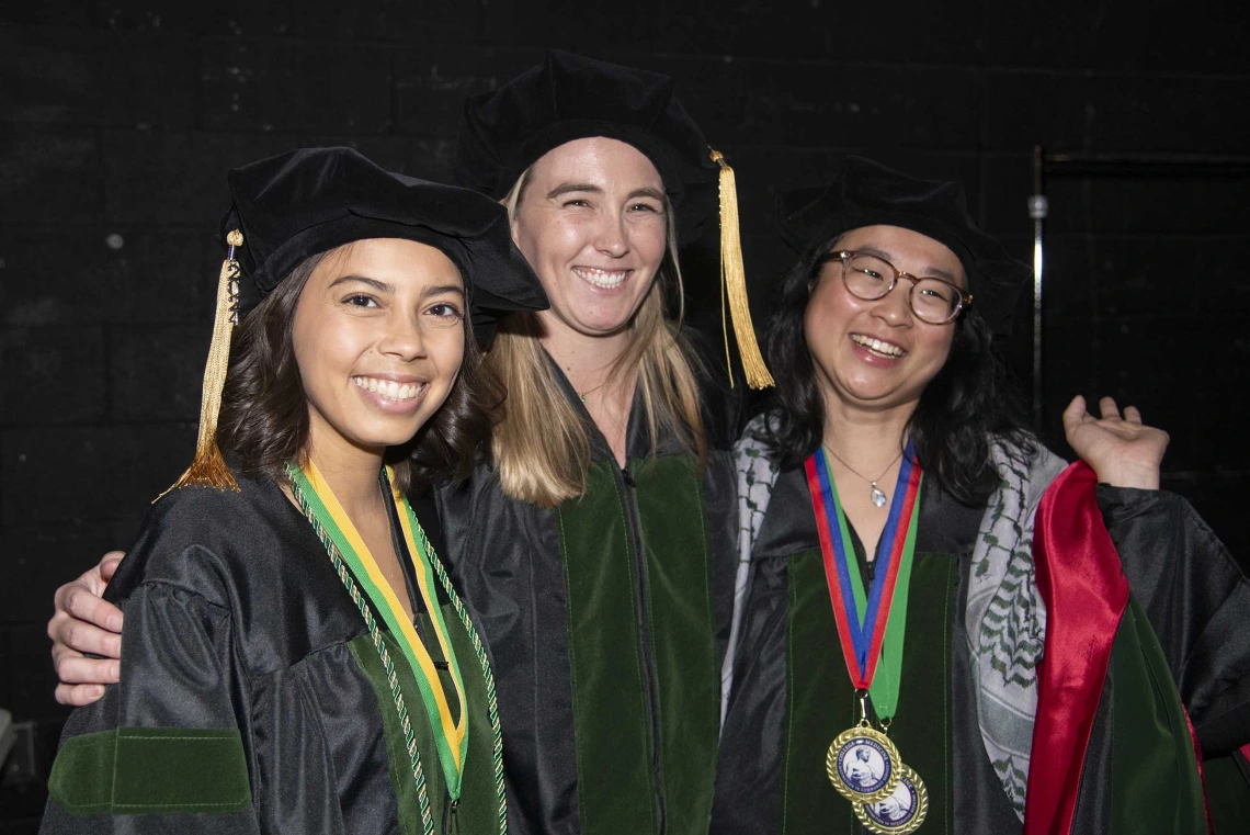 Three University of Arizona College of Medicine – Tucson students in graduation caps and gowns stand together, smiling.  