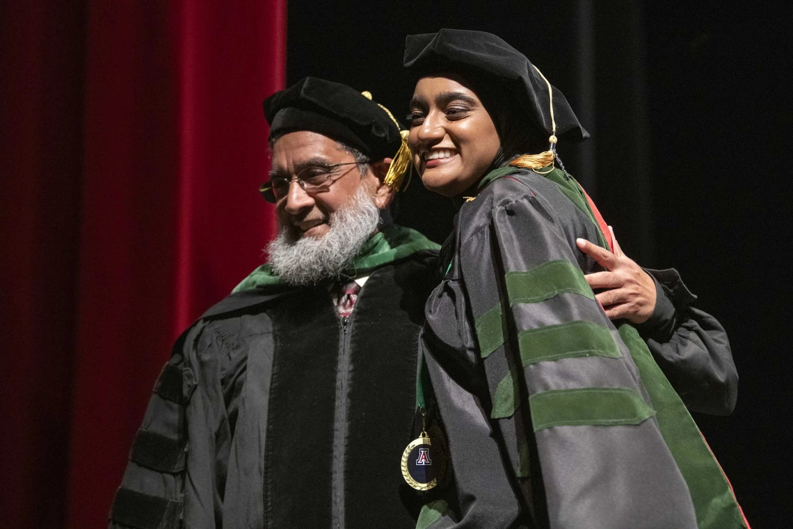 A University of Arizona College of Medicine – Tucson student dressed in a graduation cap and gown hugs her father, who is also dressed in graduation regalia, while standing on a stage.